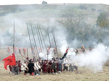 A colour photograph showing a re-enactment of a seventeenth century battle, with a unit of infantry firing muskets.