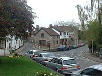Street scene showing road junction and grey stone buildings with parked cars in front of them. To the left is a grassy area with a tree.