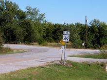 Un letrero que dice "FIN" sobre el escudo SH-49.  El letrero se encuentra junto a una carretera que cambia bruscamente de una carretera rayada bien cuidada a una carretera sin rayas de menor grado en el mismo lugar que el letrero.