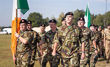 Male and female soldiers wearing camouflage marching behind the Irish tri-color flag.