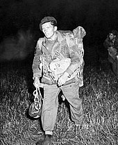 man in Brits legeruniform, met parachutehelm op en baret op, andere mannen zijn nog net te zien op de donkere achtergrond