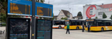 Bahnhofplatz Wil mit elektronischer Abfahrtstafel, einem Fahrzeug des Regiobus und zwei Postautos