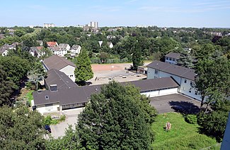 Blick von einem gegenüberliegenden Hochhaus: der Gebäudekomplex besteht aus drei senkrecht zueinander angeordneten länglichen, weißen Gebäuden mit grau gedeckten Giebeldächern; dahinter Schulhof und Sportplatz, vorn Lehrerparkplatz und Wiesengelände.