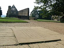 Photograph shot low to the ground near an inscribed stone set into the ground, with a background of ruined stone buildings.