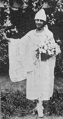 A young, smiling African woman, standing outdoors, wearing a white dress with wide sleeves, a pointed hat low across her brow, and holding a bouquet of flowers.
