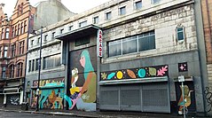 Kentsel bir caddede bir binanın gri taş Art Deco cephesi. Panjurlar zemin seviyesinde indirilmiş ve üst pencereler pencere görünümleriyle değiştirilmiştir. Zemin kat, içinde çeşitli hayvanların bulunduğu bir duvar resmi ve baykuşlu çömelmiş bir kızla boyanmıştır.