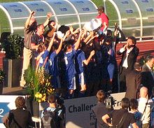 Photographie d'une équipe féminine de football, vue de profil, brandissant un trophée.