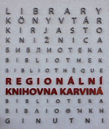 Zdjęcie przedstawia fragment ściany nowego budynku biblioteki. Na ścianie umieszczone są słowa "biblioteka" w różnych językach, jedno pod drugim. Wśród nich jest również słowo biblioteka w języku polskim. Litery są duże, drukowane, różnej wielkości - tak, aby całość tworzyła prostokąt. W jednej trzeciej wysokości jest napis w języku czeskim: regionalna biblioteka Karwina. Zajmuje on dwie linijki tekstu i wyróżnia się kontrastowym, czerwonym kolorem.