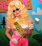 Female-presenting individual with blonde hair, colorful cosmetics, and a pink outfit with badges; she is holding marshmallows on a stick.