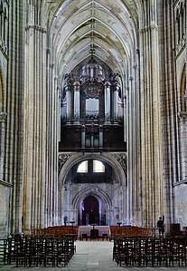 En haut de l'image, grand buffet d'orgue en bois en dessous de voûtes de pierre. En bas de l'image, portail d'église vu depuis l'intérieur, derrière des rangées de chaises en bois.