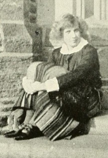 A young white woman with fair hair, seated outdoors in front of a stone building, wearing a dark top and striped skirt