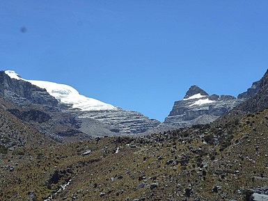 The side of Pan de Azúcar peak (left) and can be seen through a broad valley with sparse páramo vegetation. Glacially deposited boulders can be seen on the foreground and small moraines cab be observed as gray, rocky ridges in the middleground, trending in the same direction as that of the photograph. Colluvium (rock fall) deposits can be observed by the steep rocky faces to the left.