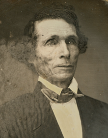 sepia-toned picture of a man with dark hair and deep-set eyes, wearing a dark suit coat, white shirt, and cravat