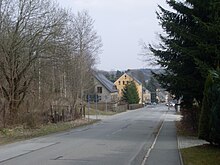 abschüssige Straße ohne Straßenbahnmarkierungen, Bürgersteig auf der rechten Seite, links und rechts Bäume, hinten ein paar Häuser, Autos und Ampel