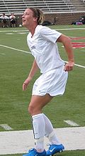 Wambach portant un uniforme blanc près d'un terrain de football.