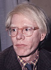 Andy Warhol, a man with light-colored hair, light skin, and glasses. He is wearing a patterned shirt and a tie under a jacket. He appears to be middle-aged and is looking slightly to the right of the camera. His expression is neutral, and he has a distinctive, pale complexion.