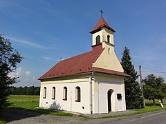 Zdjęcie wykonane w słoneczny dzień przedstawia kaplicę zwróconą frontem do drogi. Zdjęcie jest wykonane z drogi, w kierunku narożnika budynku. Kaplica jest pomalowana na biało, dach jest dwuspadowy, pokryty blachą. W ścianie frontowej umieszczone jest wejście, w bocznej znajdują się cztery okna. Nad wejściem w dachu jest niska wieża z ostrosłupowym dachem. Przy kaplicy rosną drzewa, za nią znajduje się łąka.