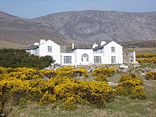 Dům Charlese Boycott na ostrově Achill.  Jedná se o velký bílý dům se dvěma podlažími.  V pozadí je vidět hornatý terén na ostrově.
