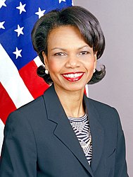 Condoleezza Rice ridetanta kun dike uzis ruĝan lipoŝminkon portantan malhelbluan jakon super strukturizita bluzo.
Usono-flago estas en la fono.