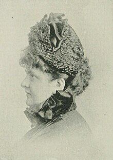 19th-century B&W portrait profile photo of a woman with her hair in an up-do, wearing a dark blouse with embellished collar, and a hat.