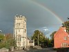 Three stage square stone church tower on the left. Red painted building on the right and a rainbow.