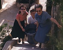 Three women carry a large tub of water up stone steps.