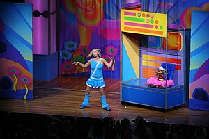 A distant stage shot of a female performer wearing bright clothes and singing into a headset microphone. A jack-in-the-box style puppet sits behind her on a movable section, which is brightly coloured.