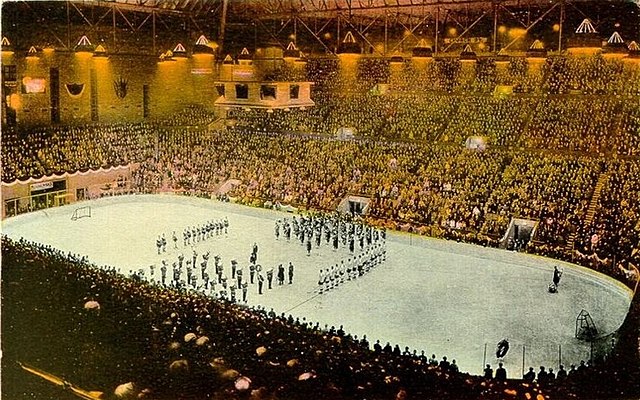 Far view of an ice hockey rink, with ice hockey players and a band standing at its centre. Spectators seated around the rink watch.