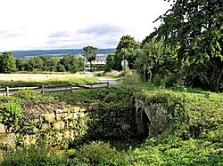 Kamenný mostek přes starou cestu ze Slotova do Kašova na jižním okraji obce Kuks, v pozadí je kukský hospitál, založený F. A. Šporkem