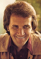A smiling brown-haired man wearing a brown jacket