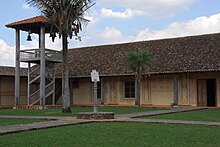 Um pátio de igreja com um relógio de sol no centro do pátio e uma torre sineira de madeira no canto do pátio.  A torre sineira é constituída por uma plataforma coberta suportada por quatro colunas de madeira.  As escadas conduzem à plataforma.  O relógio de sol está localizado no final de uma coluna de madeira.