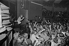 photo en noir et blanc d'un groupe de musique jouant dans une salle pleine.