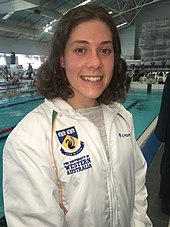 Tamsin Cook mit weißer Trainingsjacke in der Schwimmhalle. Die Trainingsjacke trägt die Aufschrift ihrer Universität.