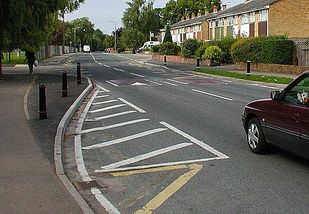 Two traffic calming measures on a road in England: speed cushions (the two reddish pads in the road) and a curb extension (marked by the black posts and white stripes)
