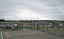 Depicts the outside of the school. Green gates encompass the building. In. the distance in front of the building is a car park. To the right, behind the gates, there is grass with a yellow digger.