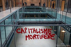 Etichetta in vernice rossa: "Capitalismo mortale" in una sala pulita e vetrata.