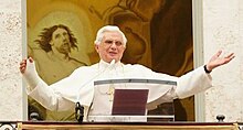Farbige Untersicht vom Papst, der auf einem Balkon mit Rednerpult steht und seine Arme ausbreitet. An seiner rechten Hand ist ein Gips. Im Hintergrund ist teilweise ein religiöses Fresko mit einem Mann zu sehen.