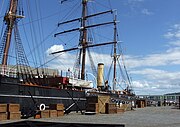 Partial view of a ship moored to a quayside. Prominent visible features are a mast with three crossbeams, two smaller masts, a funnel, a lifeboat and rigging. Packing cases are lined up on the quay, and a gangplank with "RRS Discovery" on it leads to the ship.