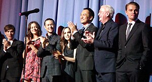 A photograph of seven people on stage; except for Leonardo DiCario on the right, they are all clapping cheerfully.