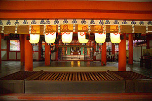 View through a hall (Haiden) with vermillion red beams and hanging lanterns to a space with standing lanterns beyond which there is another building with an altar.