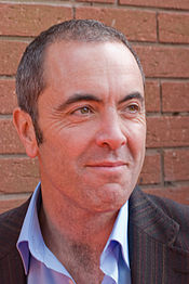 A man with a receding hairline smiles. He wears a light blue shirt with an open collar and a charcoal grey jacket.