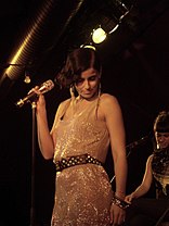 Nelly Furtado looking down while performing in a white sequin dress with a rhinestone-covered black belt.