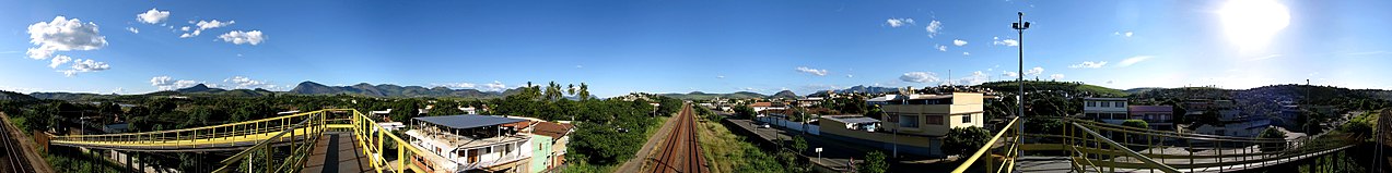 Sobre uma passarela de ferrovia se vê num dia claro de sol montes ao fundo uma pequena cidade em meio ao verde de árvores e campinas cortadas pelos trilhos da estrada de ferro.