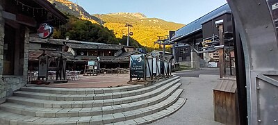 Photographie d'une placette où il y a une gare de télécabine, des commerces et des bureaux sous forme de chalets, ayant des pancartes publicitaires, le tour au pied des montagnes.