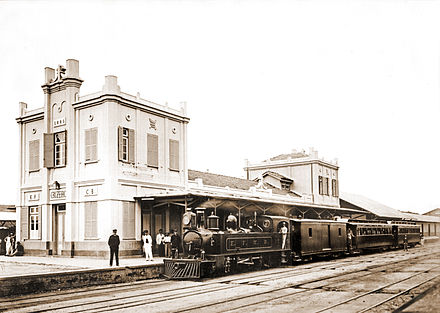 Photograph of a steam locomotive pulling passenger cars and sitting at the platform outside a large station building