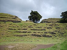 Una serie de terrazas de piedra seca semi-derrumbadas, cubiertas de hierba corta.  En la parte superior de la más alta de las cinco terrazas se encuentran los restos desmoronados y cubiertos de maleza de dos grandes edificios que flanquean las ruinas de una estructura más pequeña.  Un árbol crece del lado derecho del edificio central más pequeño, y otro se encuentra en el extremo derecho, en la terraza superior y frente al edificio también de pie sobre él.  El primer plano es un área de plaza plana, con el flanco derrumbado de una pirámide cubierta de hierba en la parte inferior derecha.  El cielo está nublado con nubes de lluvia bajas.