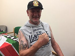 Man wearing sleeveless hoodie with tattoos on right arm and right fist clenched