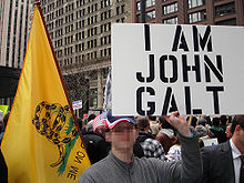 In a large outdoor crowd, a man holds up a poster with the words "I am John Galt" in all capital letters