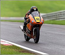 Moto de corrida com carroçaria marrom e o piloto dobrado em uma pista lisa com um parque verde ao fundo