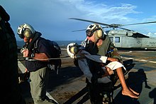 In the background, a helicopter. In the foreground, two men in flight helmets, one carrying a child, the other leading.